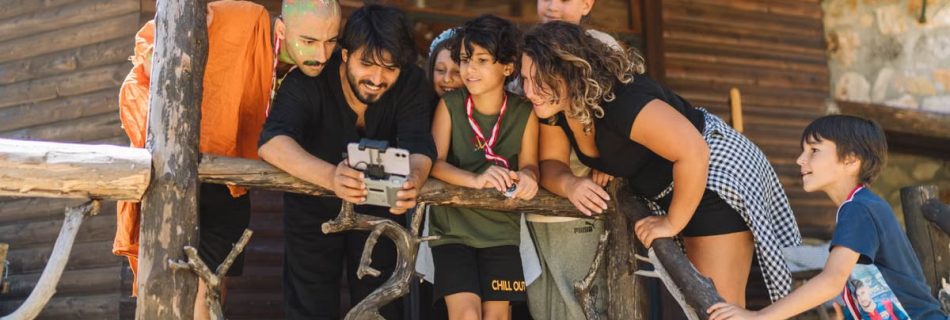 A diverse group of people joyfully watching drone footage on a wooden cabin porch.